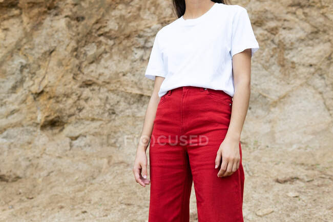 Cropped unrecognizable female adolescent in white t shirt and red jeans standing on rough land against mount — Stock Photo