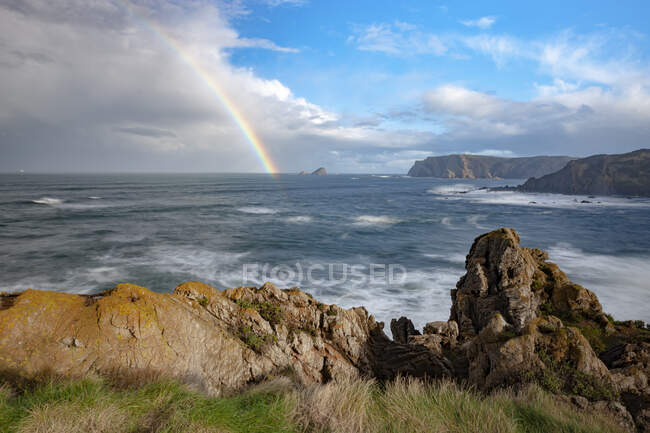 Tolle Landschaft mit Regenbogen und Wolken am blauen Himmel über welligem stürmischem Meer, das die felsige Küste in Verdicio Asturias spült — Stockfoto