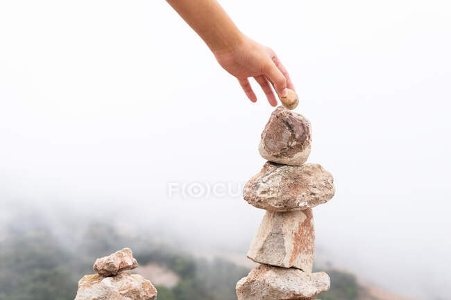 Crop anonymous female tourist placing stone in pile while exploring mountainous terrain covered with fog — Stock Photo