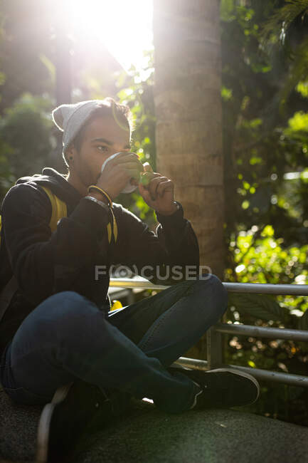Friedlicher afroamerikanischer Hipster mit Rucksack chillt im Park und trinkt Erfrischungsgetränk to go — Stockfoto