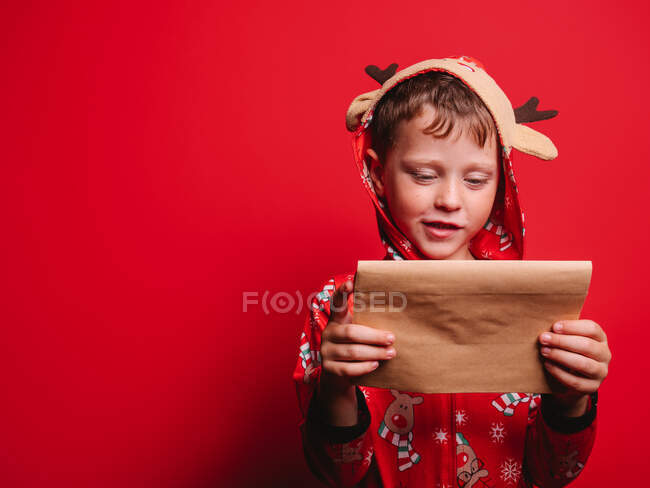 Happy little boy festive costume reading letter against red background during Christmas party — Stock Photo