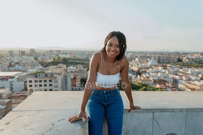 Content young African American female in jeans and crop top looking at camera on fence in city — Stock Photo