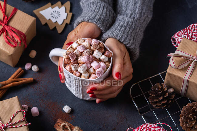 Von oben hält eine Frau einen Becher Heißgetränk mit Marshmallows zwischen Weihnachtsgeschenken und Zimtstangen — Stockfoto