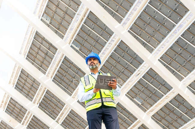Niedriger Winkel eines hispanischen Ingenieurs mittleren Alters in Uniform mit Tablet, der unter einem Solarkraftwerk wegschaut — Stockfoto