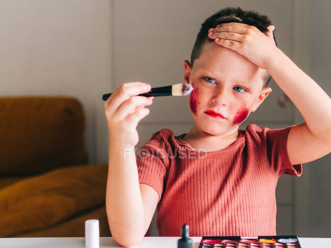 Charming child with makeup applicator touching head while looking away at table with eyeshadow palette — Stock Photo