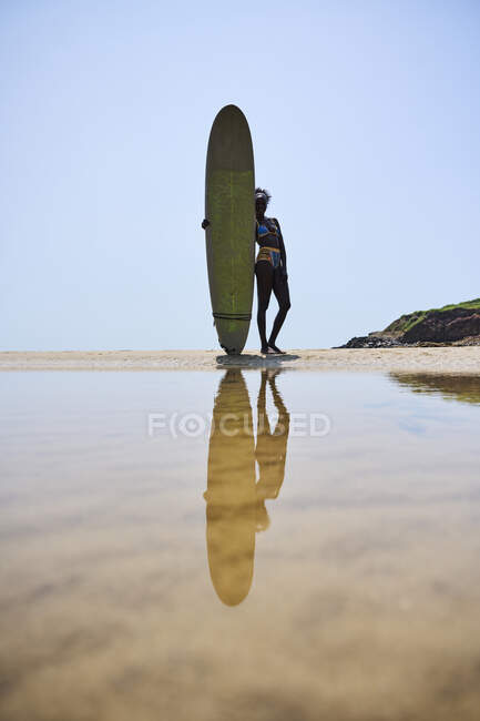 Back view of African American female athlete with surfboard admiring ocean from sandy shore under cloudy blue sky — Stock Photo