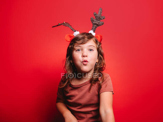 Niña alegre en ropa casual y diadema de ciervo festivo soplando beso mirando a la cámara durante la celebración de Navidad contra el fondo rojo - foto de stock