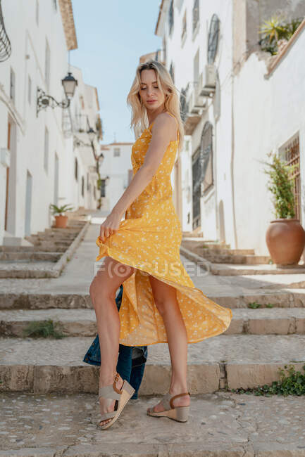 Full body side view of young female in summer outfit standing on stone stairway between old buildings in alley — Stock Photo