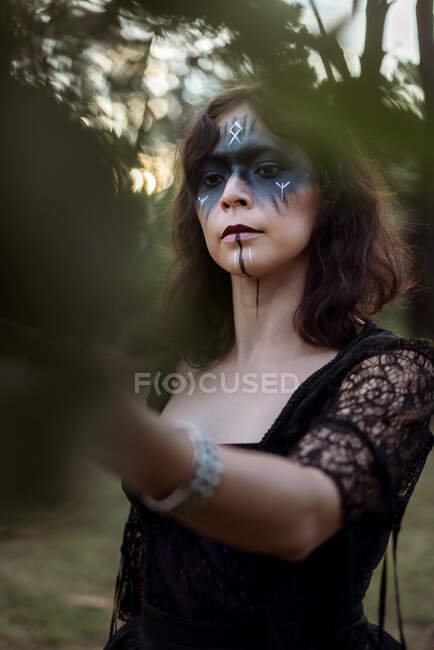 Mystic witch in long black dress and with painted face standing looking and touching leaves in dark gloomy woods — Stock Photo