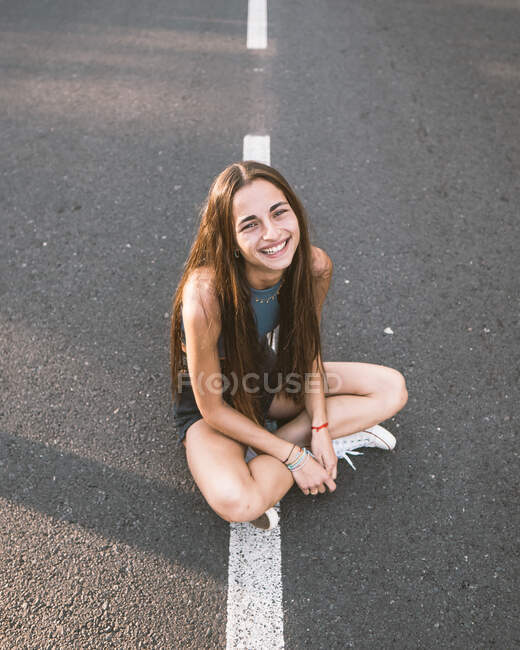 From above of friendly female adolescent with long hair sitting on asphalt roadway while looking at camera in Tenerife Spain — Stock Photo