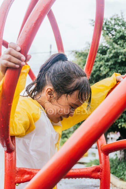 Seitenansicht von fröhlichen ethnischen Kind mit nassen Haaren in Slicker Spaß zwischen Metallkonstruktion bei regnerischem Wetter — Stockfoto
