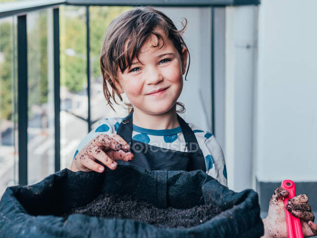 Positive little girl in apron with flower pot and playing dirty hands on balcony in daytime while looking at camera — Stock Photo