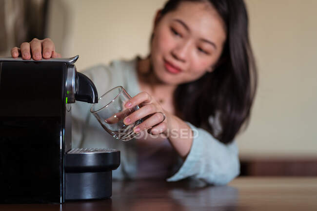 Young ethnic female against pod coffee maker pouring hot beverage with foam into glass in house kitchen — Stock Photo