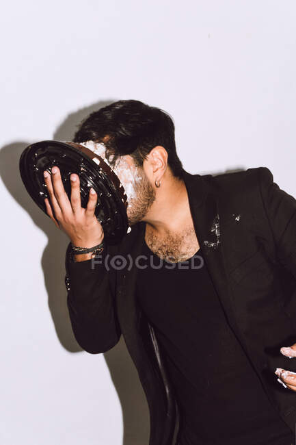 Unshaven unrecognizable male in black outfit smashing birthday cake in face during festive party in studio — Stock Photo