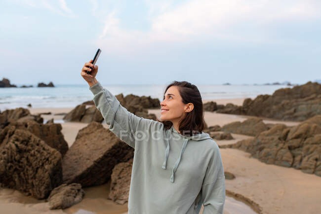 Friendly female taking self portrait on cellphone on sandy shore against rocks and ocean under cloudy sky in Asturias Spain — Stock Photo