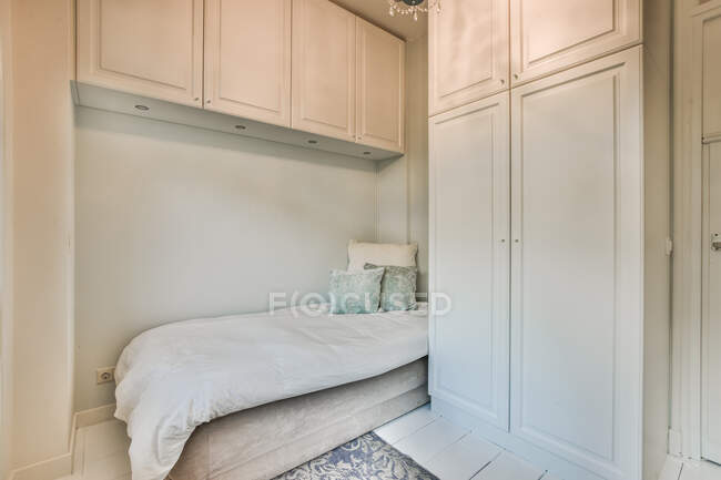 Комфортабельная кровать с одеялом в минималистском стиле в спальне с белым столом и шкафами в современной квартире — стоковое фото
