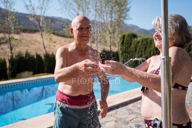 Чоловік без сорочки стоїть біля старої позитивної дружини і приймає душ у басейні в сонячний літній день на задньому дворі — стокове фото