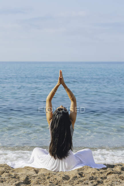 Back view of anonymous graceful female with long hair and namaste hands sitting on sandy beach during yoga practice against ocean — Stock Photo