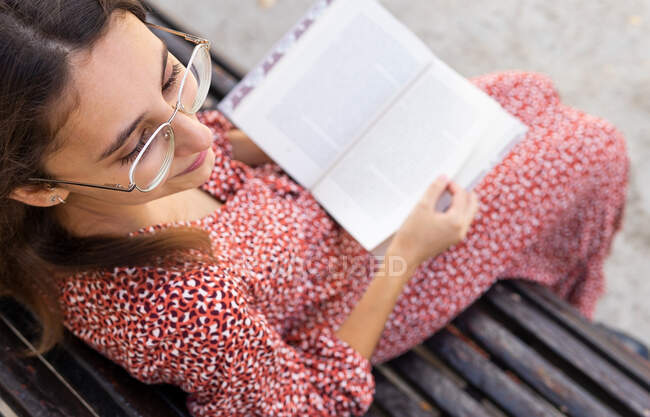 From above positive young female in stylish clothes sitting with opened book on wooden bench against building with light wall in daytime — Stock Photo
