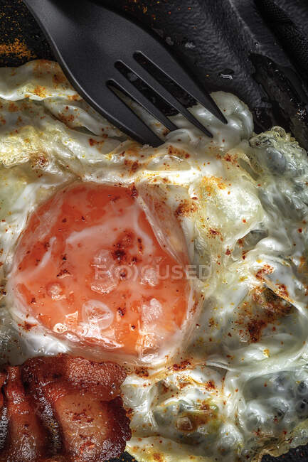 From above of sunny side up egg with fried bacon slices and condiments on tray against cutlery on dark background — Stock Photo