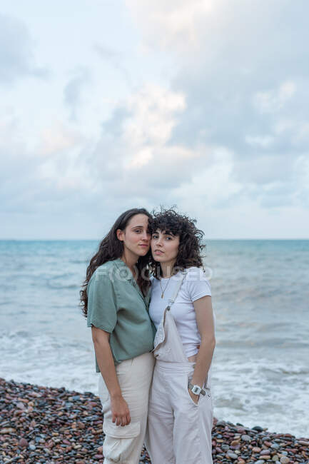 Девушки-лесбиянки в повседневной одежде обнимаются, глядя в камеру на побережье океана под облачным небом — стоковое фото