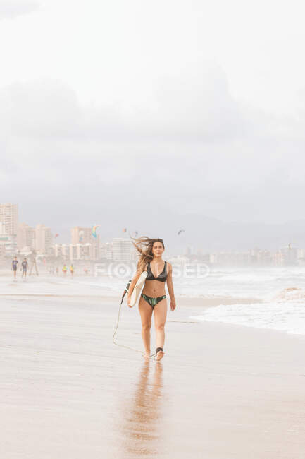 Joven atleta en traje de baño con pelo volador y tabla de surf caminando y mirando a la cámara en la costa del océano - foto de stock