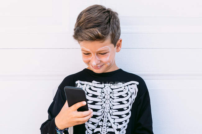 Menino alegre em traje de esqueleto preto com rosto pintado usando no telefone celular enquanto sentado perto da parede branca na rua — Fotografia de Stock