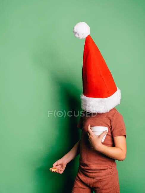 Ragazzo irriconoscibile che indossa il cappello rosso di Babbo Natale per festeggiare il Natale e mangia dolce biscotto sullo sfondo verde — Foto stock