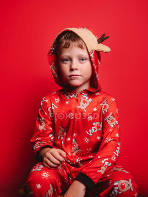 Unbeweglicher süßer kleiner Junge im Kapuzenpyjama mit Hirsch, der im roten Studio in die Kamera schaut — Stockfoto