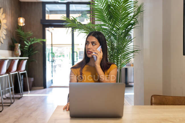 Freelancer feminino sério sentado à mesa de madeira no café e ter uma chamada telefônica enquanto digita no netbook olhando para longe — Fotografia de Stock