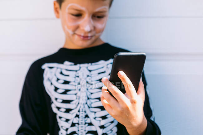 Cheerful boy in black skeleton costume with painted face using on mobile phone while sitting near white wall on street — Stock Photo
