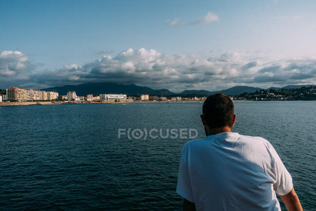 Обратный вид анонимного туриста-мужчины, созерцающего бурлящий океан и сидящего под кучевыми облаками в Сен-Жан-де-Луз — стоковое фото