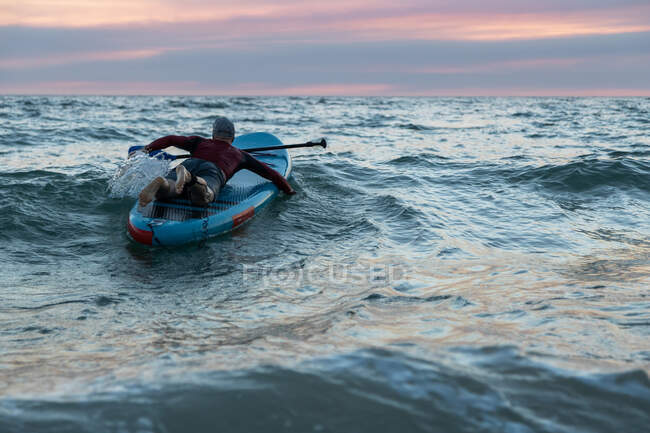 Back view of unrecognizable male surfer in wetsuit and hat on paddle board surfing on seashore — Stock Photo