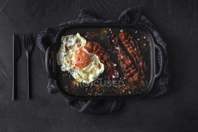 Vista superior do lado ensolarado ovo com fatias de bacon frito e condimentos na bandeja contra talheres no fundo escuro — Fotografia de Stock