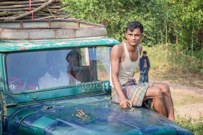 INDIA, BANGLADESH - DECEMBER 5, 2015: Young ethnic male in casual clothes sitting on a old car in rural terrain near green trees and looking at camera — Stock Photo