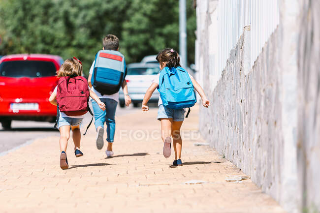 Back view of anonymous schoolkids with backpacks running on tiled walkway in sunny town on blurred background — Stock Photo