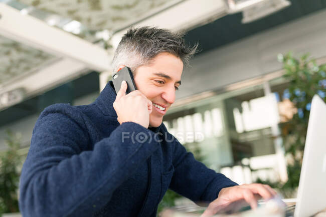Vista lateral del joven freelancer masculino en ropa casual y anteojos hablando por teléfono móvil y mirando a la cámara mientras está sentado en la mesa con portátil y portátil - foto de stock
