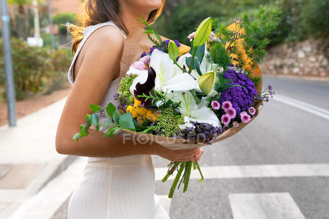 Vue latérale d'une femelle récoltée méconnaissable dans des verres avec un bouquet floral en fleurs regardant loin sur la route de passage de la ville — Photo de stock