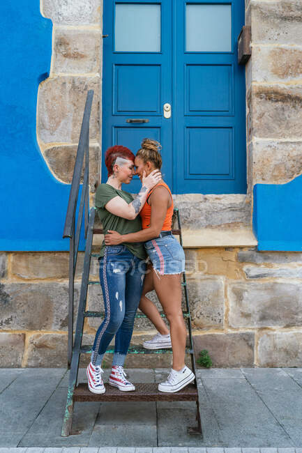 Side view of cool smiling young homosexual girlfriends embracing while touching noses on staircase against building in town — Stock Photo