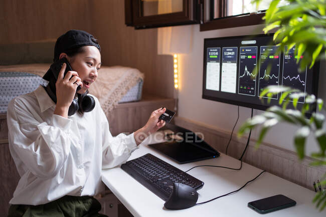 Überraschte asiatische Händlerin spricht auf Smartphone und betrachtet Kreditkarte, während sie mit Kryptowährungsstatistik arbeitet — Stockfoto