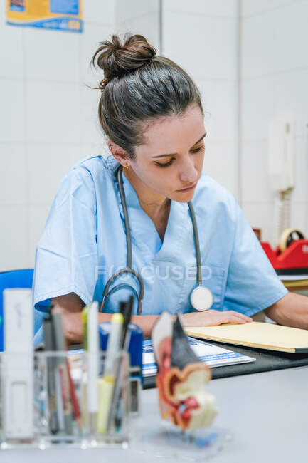 Молодая женщина-ветеринар со стетоскопом и бумажными простынями работает за столом в больнице на размытом фоне — стоковое фото