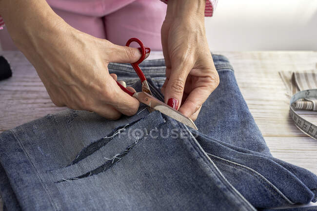 Cosecha costurera femenina anónima usando tijeras mientras se cortan pantalones vaqueros en la mesa de madera en el taller - foto de stock