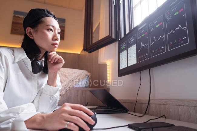 Visão lateral da mulher asiática concentrada trabalhando no computador com gráficos mostrando dinâmica de mudanças no valor da criptomoeda no local de trabalho conveniente — Fotografia de Stock