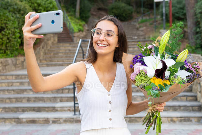 Content young female in eyeglasses with blossoming flower bouquet taking selfie on cellphone on urban stairs — Stock Photo