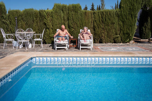 Coppia anziana spensierata in costume da bagno e occhiali da sole prendere il sole su lettini con bere caffè vicino alla piscina e siepe — Foto stock