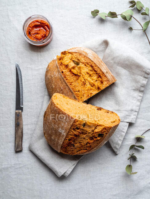 Top view of freshly baked loaf of bread and red pesto sauce in glass jar served on table with napkin and knife — Stock Photo
