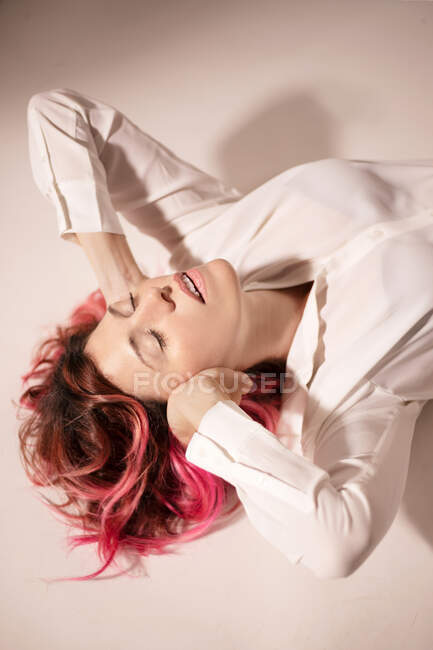 Зверху спокійна жінка з рожевим волоссям, що лежить з закритими очима на підлозі і покриває вуха руками в світлій кімнаті — стокове фото