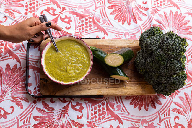 Vista dall'alto del raccolto anonimo femminile con cucchiaio in ciotola con purea di zucchine e broccoli su tagliere di legno — Foto stock