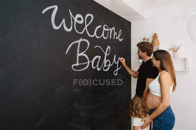 Homme écriture Bienvenue Bébé inscription sur tableau noir contre femme enceinte bien-aimée et fille à la maison — Photo de stock