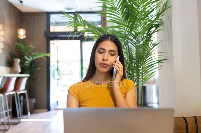 Serious female freelancer sitting at wooden table in cafe and having phone call while typing on netbook — Stock Photo
